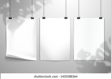 The shadow of the Monstera plant falls on three white blank realistic sheets of paper hangs on the clamps. Mockup for design