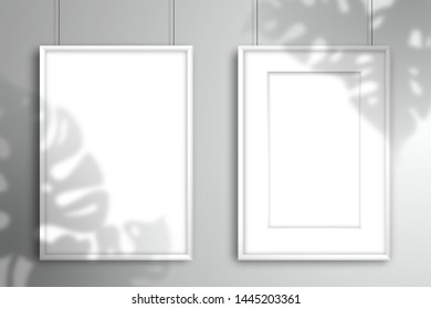 The shadow of the Monstera plant falls on a two empty white frames with passe-partout and without it for an image or photo. Mockup for design