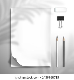 The shadow of a palm leaf falls on a notebook with a bent top sheet, two pencils, binder and eraser. Flat lay with blank white elements for design