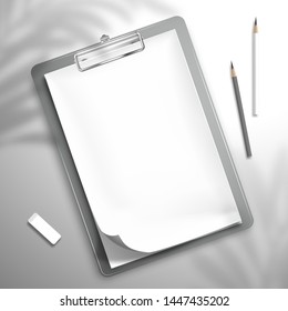 The shadow of a palm leaf falls on a white blank notebook with a bent top sheet is clamped in the clipboard, two pencils and an eraser that are scattered over the surface. Top view mockup template