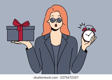 Shocked woman with gift and alarm clock in hands opens mouth in surprise after learning about being late for party. Businesswoman in formal black clothes and sunglasses holding gift for boyfriend