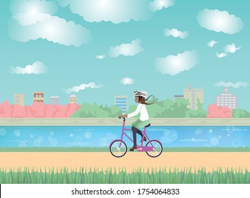 Side view of a woman riding a bicycle and wearing a helmet on a trail. Beautiful lake surrounded by a grove of trees, park and buildings. Blue sky with clouds. Calm and peaceful summer day.