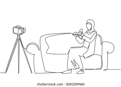 Single continuous line drawing Arabian beauty blogger is sitting at sofa, reviewing heels shoe in her hands while recording video with digital camera, tripod. One line draw design vector illustration