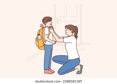 Single mother prepares young son of student in elementary school as trip to lessons and helps to put on briefcase kneeling down. Caring mother dresses teen boy for first day of school 