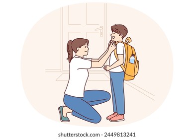 Single mother prepares young son of student in elementary school as trip to lessons and helps to put on briefcase kneeling down. Caring mother dresses teen boy for first day of school