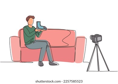 Single one line drawing social media influencer reviewing boots. Smiling young man vlogging about men's sports shoe and filming himself at home on a video camera. Continuous line draw design vector