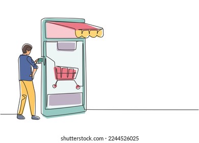 Single one line drawing young man inserting credit card into large canopy smartphone screen with shopping cart. Digital payment concept. Modern continuous line draw design graphic vector illustration