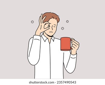 Sleepy man with mug of coffee, opens eyes with hand and needs to rest due to lack of energy and drowsiness. Tired young guy drinks coffee drink to cheer up and continue productive work.