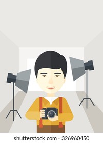 A smiling Asian photographer holding a camera and standing inside the studio with the light vector flat design illustration. Vertical layout with a text space.