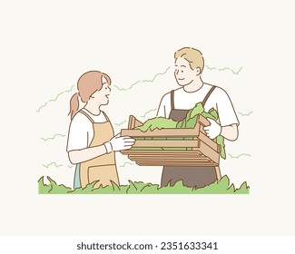 Smiling attractive young  farmer couple in apron standing, holding a wooden crate full of a variety of organic vegetables freshly harvested from an organic greenhouse farm. 