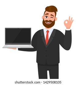 Smiling business man holding or showing a new brand, blank screen laptop computer (PC) and gesturing, making okay, OK, cool sign while winking eye. Modern lifestyle, digital technology device, gadget.