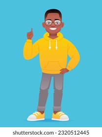 A smiling little black boy stands in glasses and points his finger at something. Cute african school kid in yellow hoodie. Vector cartoon illustration isolated on blue background.