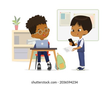 Smiling multinational primary classmates talking at classroom vector flat illustration. Happy schoolboy wearing school uniform use laptop, smartphone and paper sheet with task at lesson class interior