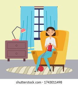 Smiling young woman knitting red scarf sitting in armchair at home. Chest of drawers with lamp, window with curtains decorating room. Quarantine isolation at home. Hobby and leisure activities