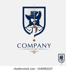 A sophisticated mascot logo featuring a classic musketeer with a signature wide-brimmed feathered hat and a full beard. The design centers on a swordsman holding a rapier vertically, integrated into a protective shield emblem with sunburst accents. This professional vector illustration uses a navy blue and bronze color palette, making it ideal for fencing clubs, historical gaming clans, heritage branding, and prestige security firms.