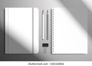 A spiral diary, a notebook with an elastic band, binder and two pencils. Flat lay mockup with shadow from window