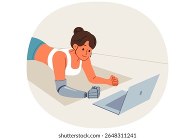 Sports woman with prosthetic arm trains at home and watches video lesson on laptop, lying on floor in plank. Sports girl doing yoga on fitness mat, training to improve health and muscle development
