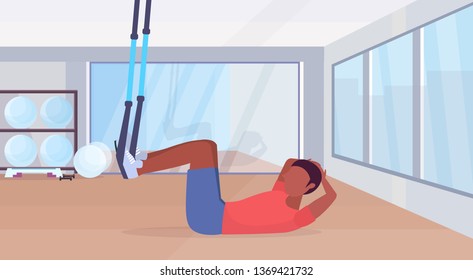 sporty man doing sit-ups abdominal exercises with suspension fitness straps elastic rope african american guy training  workout concept gym studio interior full length