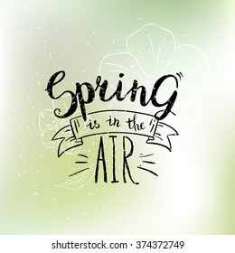 Spring is in the air card. Hand drawn calligraphy phrase on abstract blurred background with  flowers. Pastel stylish spring backdrop
