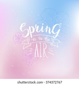 Spring is in the air card. Hand drawn calligraphy phrase on abstract blurred background with  flowers. Pastel stylish spring backdrop