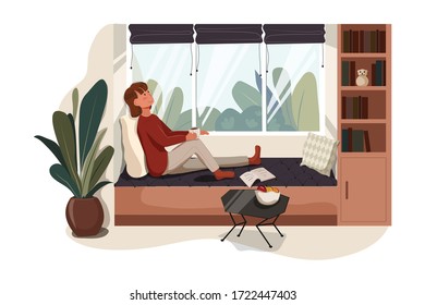 Stay at home concept. Young woman at home sitting near window relaxing in her living room reading book and drinking coffee or tea.