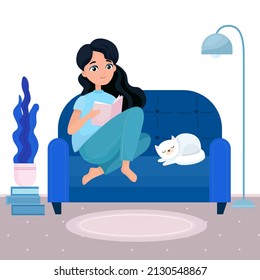 Stylish girl sitting and reading book on blue sofa with cat at home