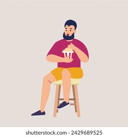 Stylish guy with a beard sits on a chair and eats popcorn on a white background
