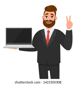 Successful business man holding or showing a new brand laptop computer (PC) and gesturing, making victory, V, peace or two sign with hand fingers. Modern lifestyle, digital technology device, gadget.
