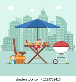 Summer picnic table on city background. Family barbecue concept with picnic party stuff. Bbq grill, straw basket and food for outing on public park.