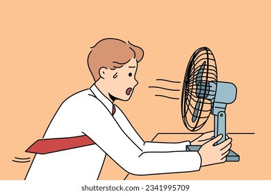 Sweaty man sits in front of fan enjoying cold wind from electric climate control equipment. Young businessman with fan suffers from lack of air conditioning and ventilation in office