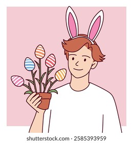 A teenage boy wearing bunny ears holds a plant with Easter egg-shaped decorations, smiling happily.