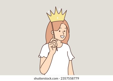 Teenage girl puts fake crown to head and smiles, dreaming of becoming princess or queen and living in castle. Happy child with golden crown on stick, participates in photo shoot during party
