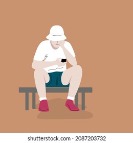 A teenager in shorts and a shirt and a Panama hat sits bent over and looks at a smartphone. The guy is fascinated by the gadget and does not notice anything around. New generation. 