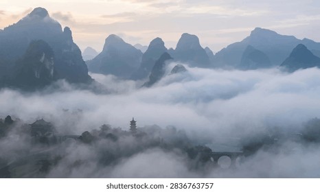 This breathtaking panoramic view captures a series of jagged mountain peaks rising above a thick, rolling blanket of morning fog. Nestled within the valley, traditional architectural silhouettes and a stone bridge are partially obscured by the ethereal mist. The soft, diffused light of early morning creates a tranquil and mysterious atmosphere, highlighting the layered textures of the rocky terrain and the calm, atmospheric depth of the scene