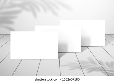 Three blank white business cards or postcards stand one after another on the plank floor. Mockup for design with palm tree shadows