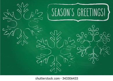 Three chalk snowflakes and a frame with hand written season's greetings over green chalkboard.