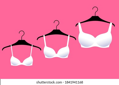 Three white brassieres with cups of different sizes are on hangers over bright pink background. Lingerie for small, medium and large female breasts. Diversity of bra sizes, fashion and beauty trends