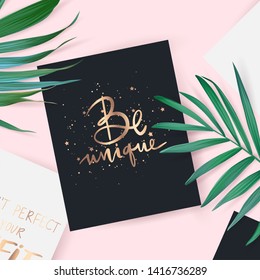 Top view with card. Be unique - Vector hand drawn lettering phrase. Motivation and inspiration gold quote. Flat lay with palm leaves