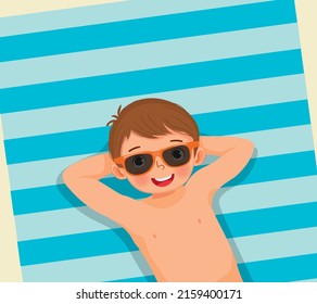 Top view of cute little boy with swimsuit and sunglasses lying on beach towel having fun sunbathing in summer vacation