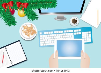 Top view of the office desk withtwig of Christmas tree. Hands are holding a tablet with blank screen ready for your text. All potential trademarks are removed.