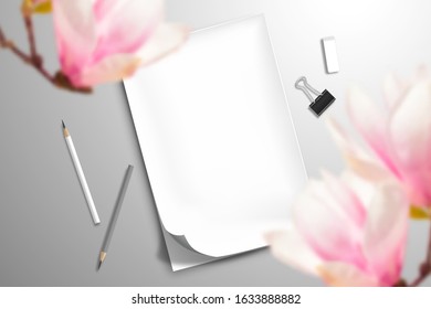 Top view on the white blank notebook with a bent top sheet, two pencils, binder and an eraser. In the foreground, large defocused magnolia flowers. Mockup template for romantic design