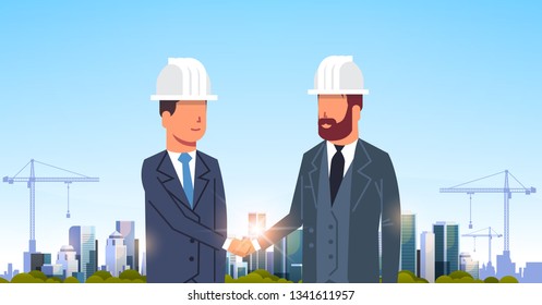 two businessmen builders handshake agreement during meeting over city construction site tower cranes building residential buildings cityscape sunset skyline background portrait horizontal