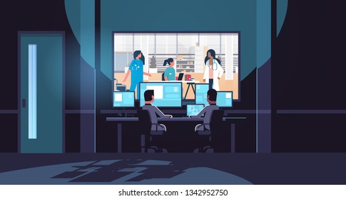 two men looking at monitors behind glass mix race doctors team in uniform discussing in medical clinic hospital dark office interior surveillance security system flat horizontal