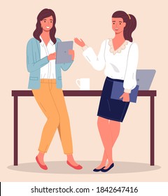 Two modern women, office workers, clerks or colleagues communicate, leaning on the table. Woman with tablet in her hand, girl with folder in her hand. Laptop and cup on the table. Coffee break