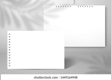 Two white blank spiral notebooks, albums or calendars. One stands on the surface, the second hangs near the wall. The shadow of the palm tree leaf falls on the wall behind. Mockup for design 