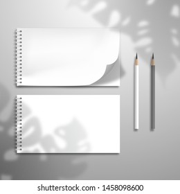 Two white blank spiral notebooks or albums and pencils. At one top sheet is bent.The shadow of the leaves of the Monstera plant is on top. Mockup for gallery design