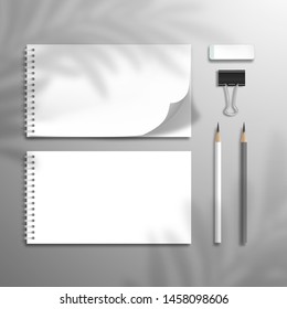 Two white blank spiral notebooks or albums. At one top sheet is bent. Lead pencils, eraser and binder. The shadow of the leaves of the palm tree is on top. Mockup for design