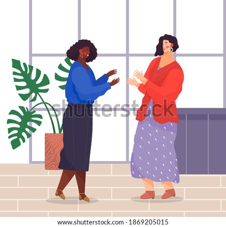 Two women standing and talking in office. Dark-skinned girl wears jewelry, blue blouse, denim skirt, white-skinned girl wears sweater, patterned dress. Women laugh and gesticulate in modern office