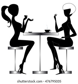 Two young chatting women sitting at a table in a modern bar, drinking coffee or tea