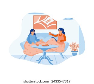 Two young women sitting on the sofa with blankets at their feet laugh, enjoying the house in winter while drinking tea with cakes. Smiling woman friends drinking tea at home. 